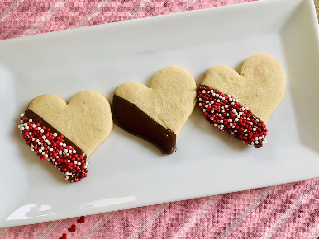 3 heart cookies on a white rectangle plate. All 3 have one edge dipped in chocolate. 2 are also dipped in red white and pink sprinkles