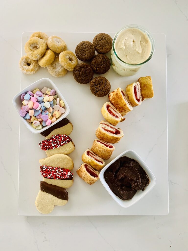 Birds Eye View: Gluten-Free Dessert Charcuterie Board with: donuts, muffins, sliced apples, cream cheese caramel dip, strawberries, candy heart, chocolate dip and Valentine sprinkled heart cookies, candy hearts, & raspberries.