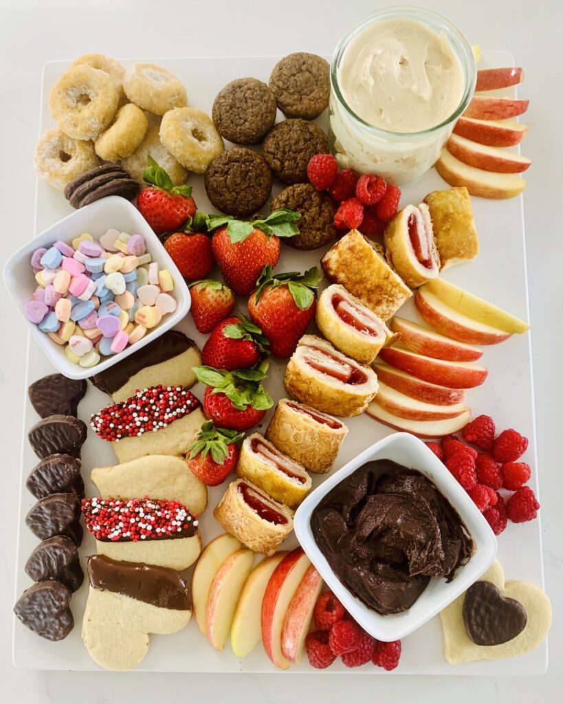 Birds Eye View: Gluten-Free Dessert Charcuterie Board with: donuts, muffins, sliced apples, cream cheese caramel dip, strawberries, candy heart, chocolate dip and Valentine sprinkled heart cookies, candy hearts, raspberries and chocolate hearts.
