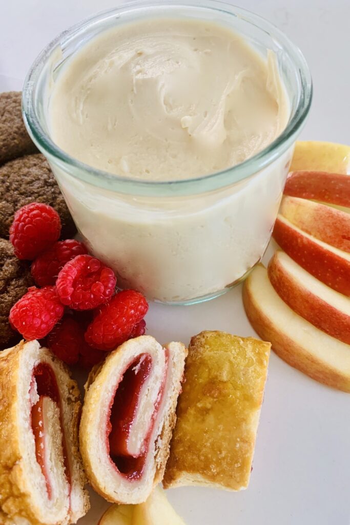 Cream cheese caramel dip in a glass surrounded by strawberry rugelach, sliced red apples, raspberries and muffins