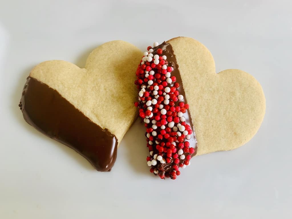 2 heart cookies with one edge dipped in chocolate. One also has been dipped in red white and pink sprinkles.