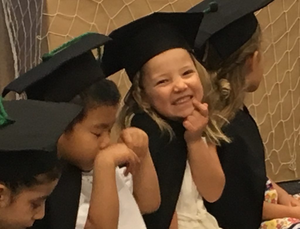 Miss E wearing a preschool graduation cap and gown and pointing to her smiling mouth