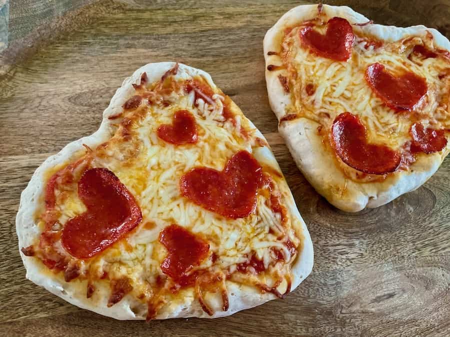 Two heart-shaped pizzas with cheese and heart-shaped pepperoni on a wood board.