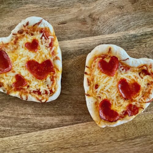 Birds Eye view: Two gluten-free heart-shaped pizzas with cheese and heart-shaped pepperoni on a wood board.