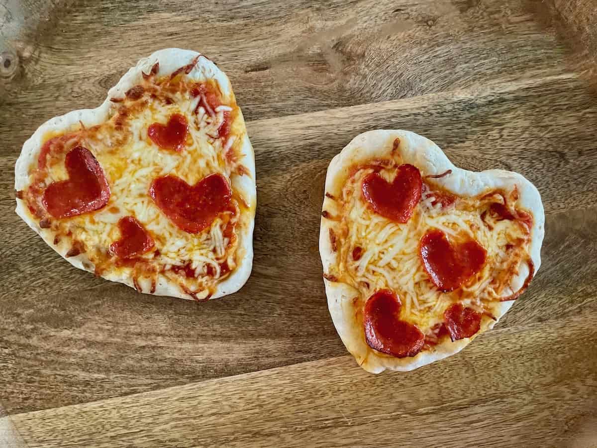 Birds Eye view: Two gluten-free heart-shaped pizzas with cheese and heart-shaped pepperoni on a wood board.