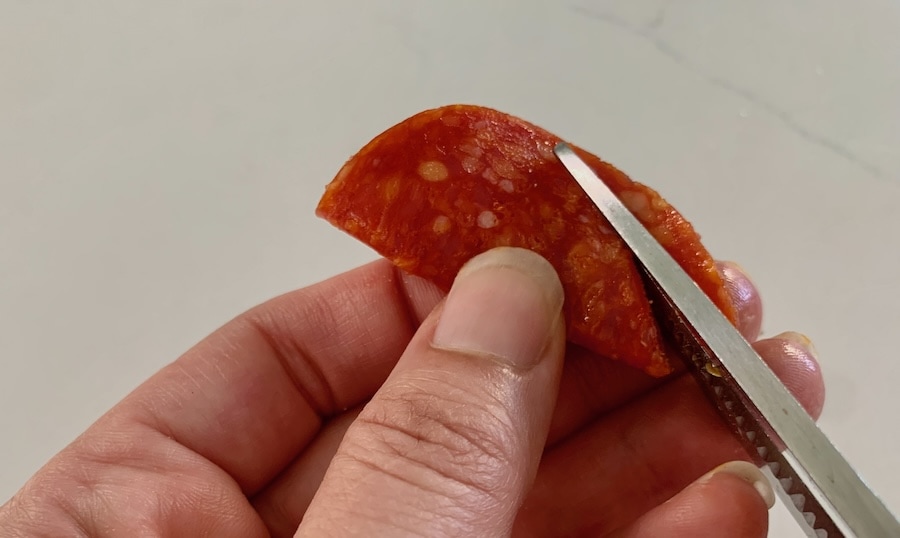 Scissors cutting a heart shape out of a slice of pepperoni folded in half.
