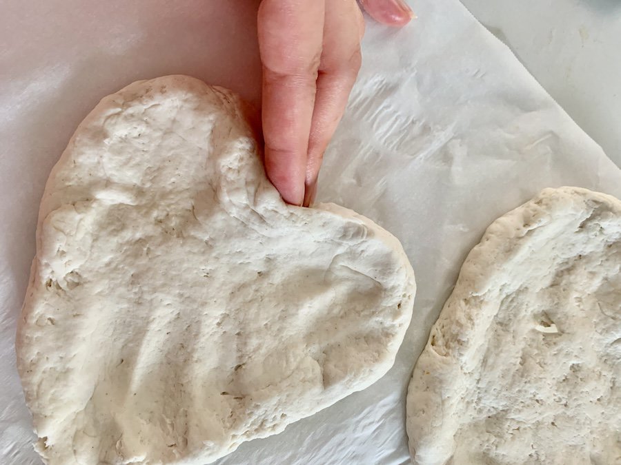 Two heart-shaped pizza crusts, unbaked. Fingers forming the heart shape top of one of the crusts.