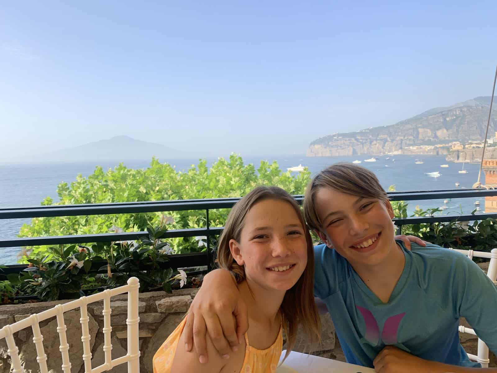 Miss E & CJ smiling at a gluten-free Italy restaurant table with a view of the bay of Naples in the background
