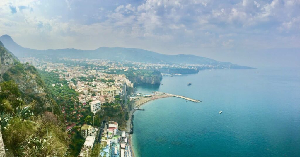 view of the turquoise bay of Naples on the right and cliffs of Sorrento and buildings on the left, puffy clouds in the sky adding a bit of haze