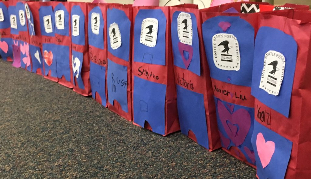 a row of red lunch bags with blue construction paper, made to look like mailboxes, each with a child's name, to be used to collect Valentine's from classmates