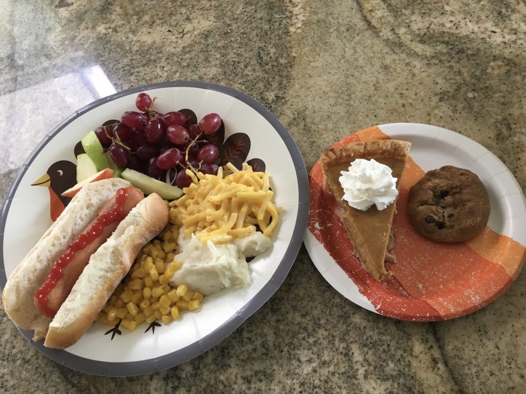Plate with gluten-free food: turkey hot dog in bun, corn, mashed potatoes, macaroni & cheese, grapes, and apple slices, and a smaller paper plate on the right with a slice of pumpkin pie and a chocolate chip cookie