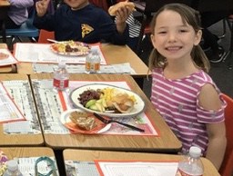 Miss E smiling at her school desk with a plate of gluten-free thanksgiving food and smaller plate of desserts in front of her on the desk