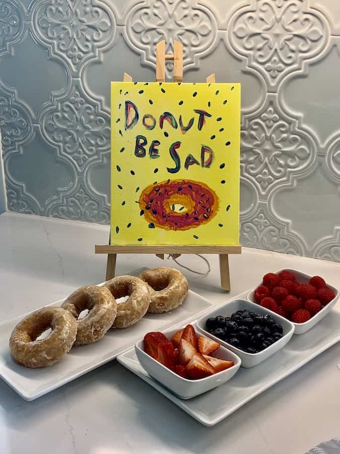 Yellow "Donut Be Sad" painting on an easel. Four gluten-free donuts with a glaze, and bowls of strawberries, blueberries and raspberries in the foreground