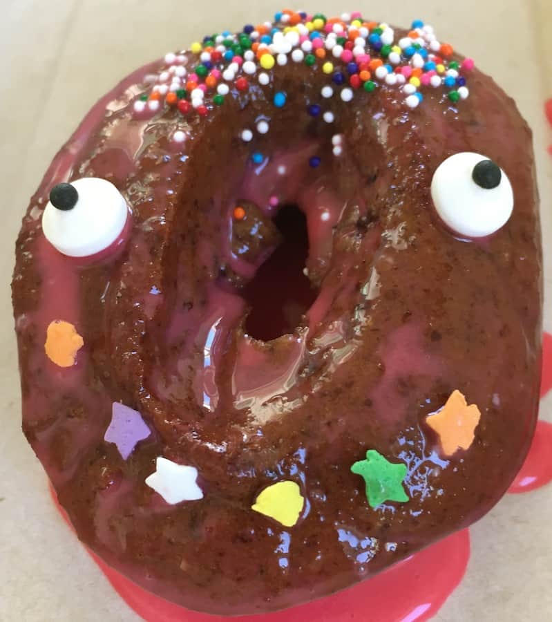 gluten free chocolate donut with strawberry glaze dripping down the side, decorations include sprinkle hair, candy eyes and star-shaped sprinkles for a mouth