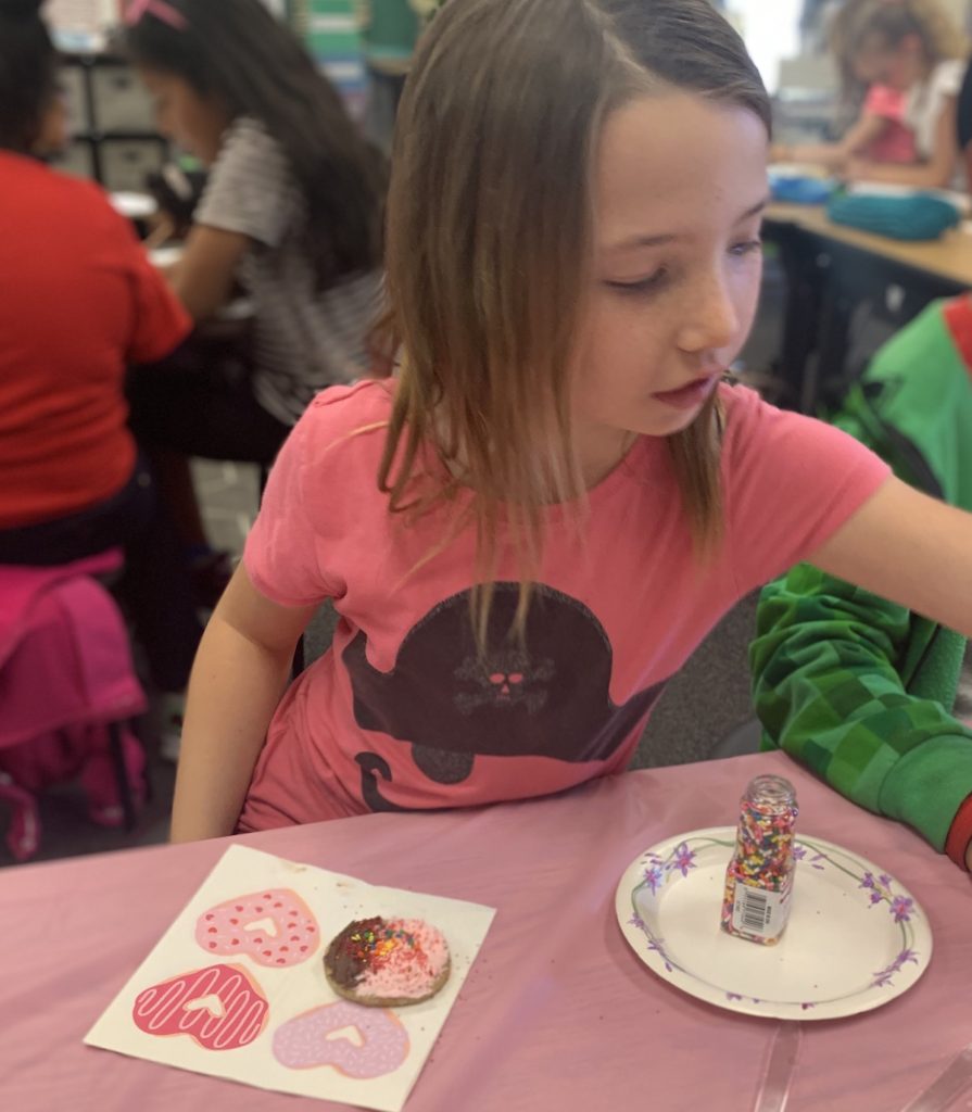 Miss E decorating a gluten-fre sugar cookie on a heart covered paper napkin, sprinkles are also on a plate in the foreground