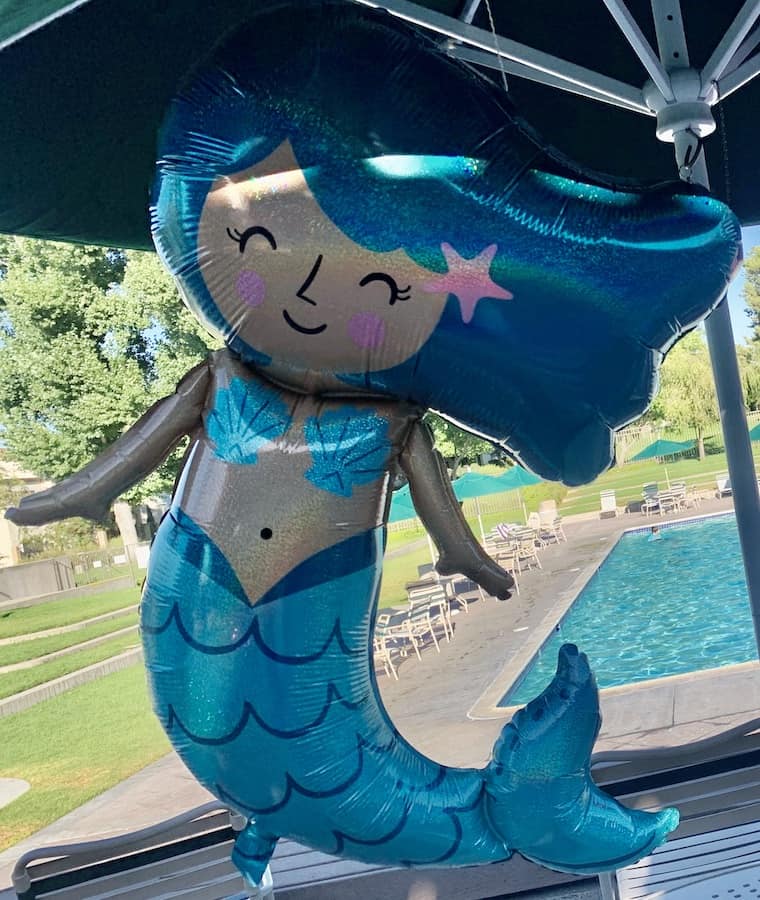 foil mermaid balloon hanging from an umbrella with pool, grass and trees in the background