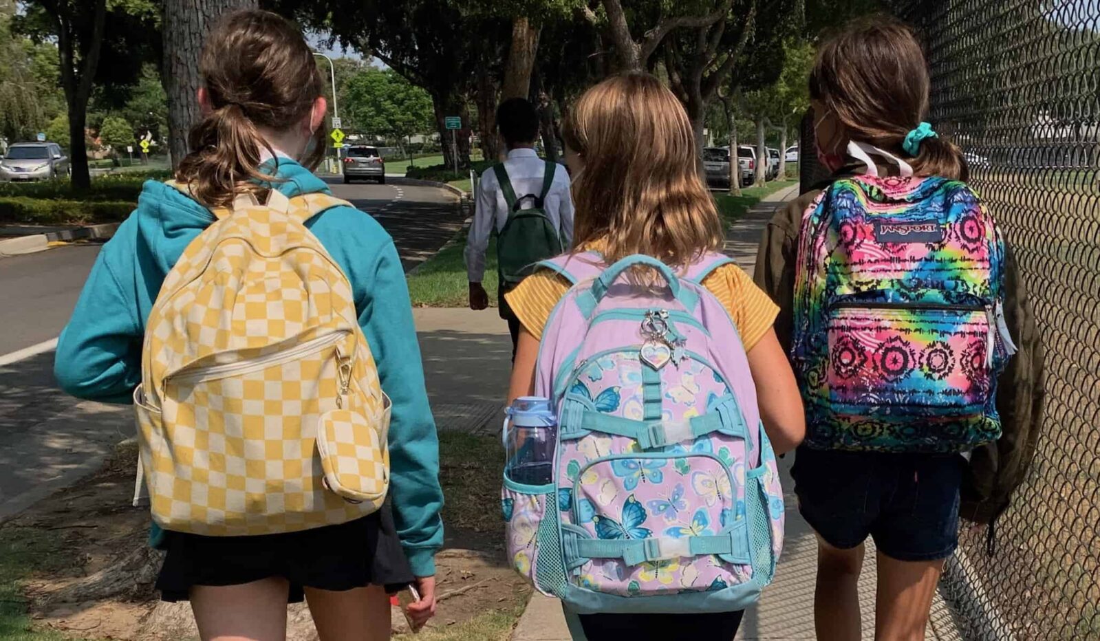 back view of three girls walking and wearing school backpacks, backpack patters are yellow checkerboard, purple and aqua butteries, and rainbow tie-dye