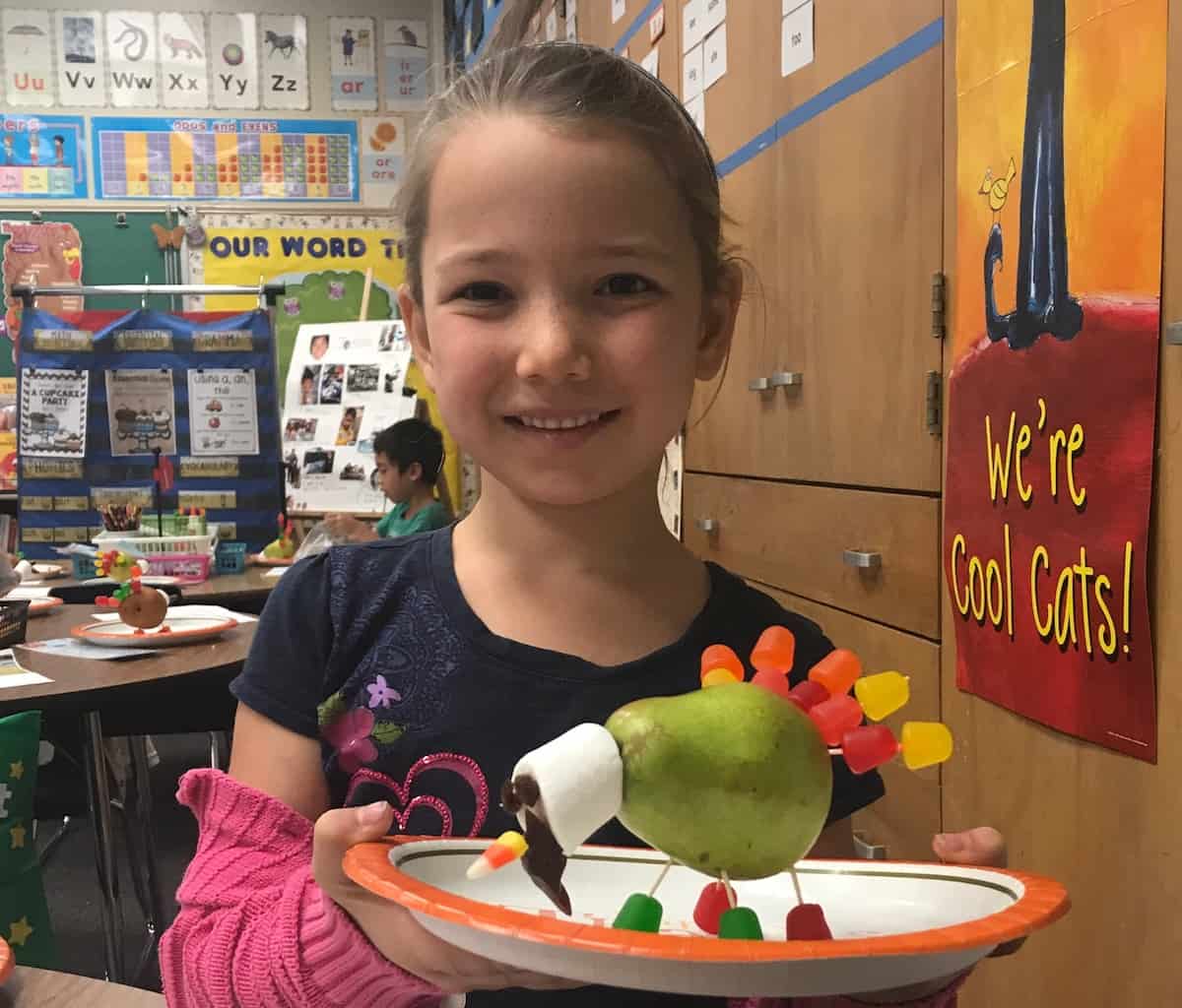 Miss E in her classroom, smiling, and holding a gluten-free turkey made of: pear, gum drop feet and feathers, toothpicks to hold candy in place, marshmallow head, raisin eyes, candy corn beak and gluten-free licorice waddle