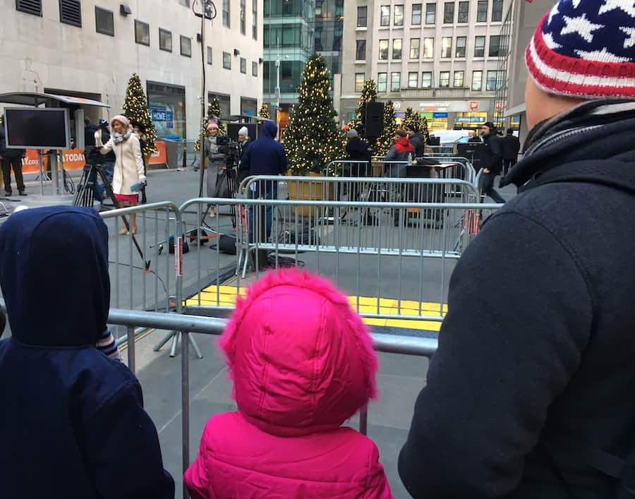 backs of family standing behind a barrier, watching Hoda Kotb, in a cream coat, along with her team walking out onto the Plaza, Christmas trees and buildings are in the background