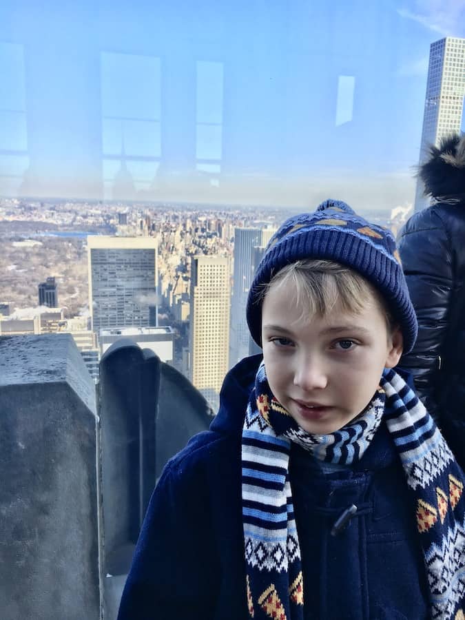 CJ wearing a hamburger/taco themed scarf and hat, half smiling on Top of the Rock with Central Park and skyscrapers in the background