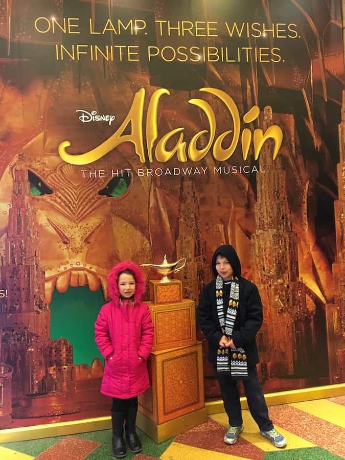 Miss E and CJ in winter coats, standing next to the magic lamp and in front of a sign that reads "Disney Aladdin" and features the Cave of Wonders with an open mouth