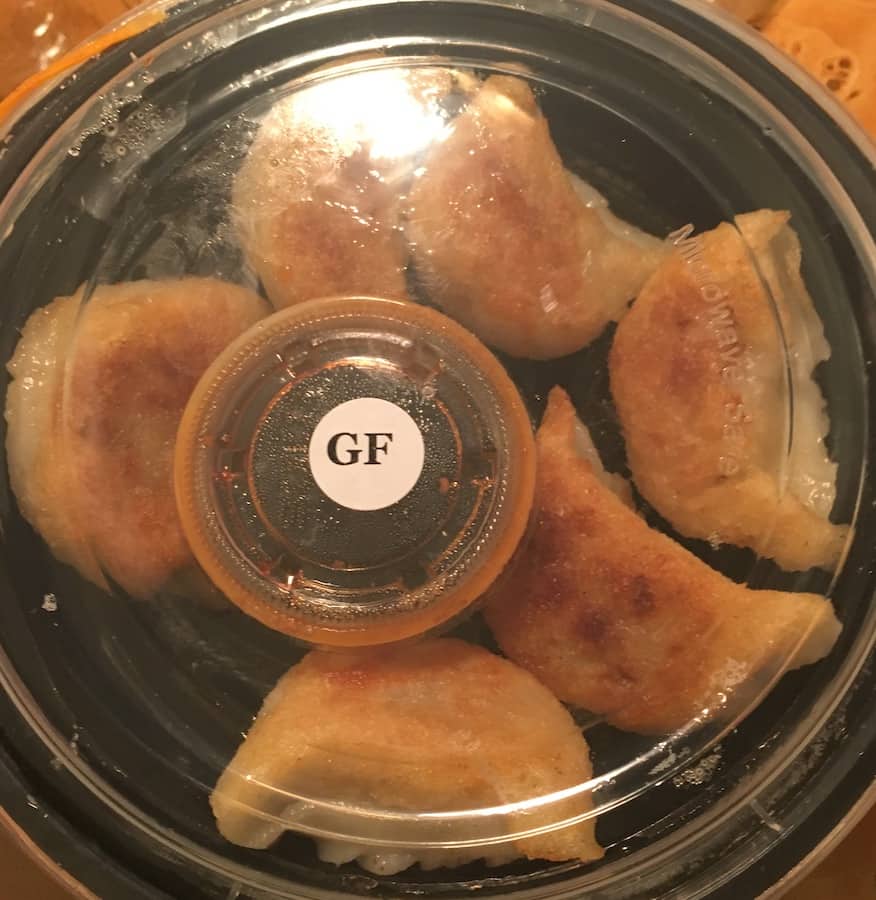 six gluten-free dumplings in a round to go container with dipping sauce in a smaller container, there is a white sticker with the letters "GF" on the container