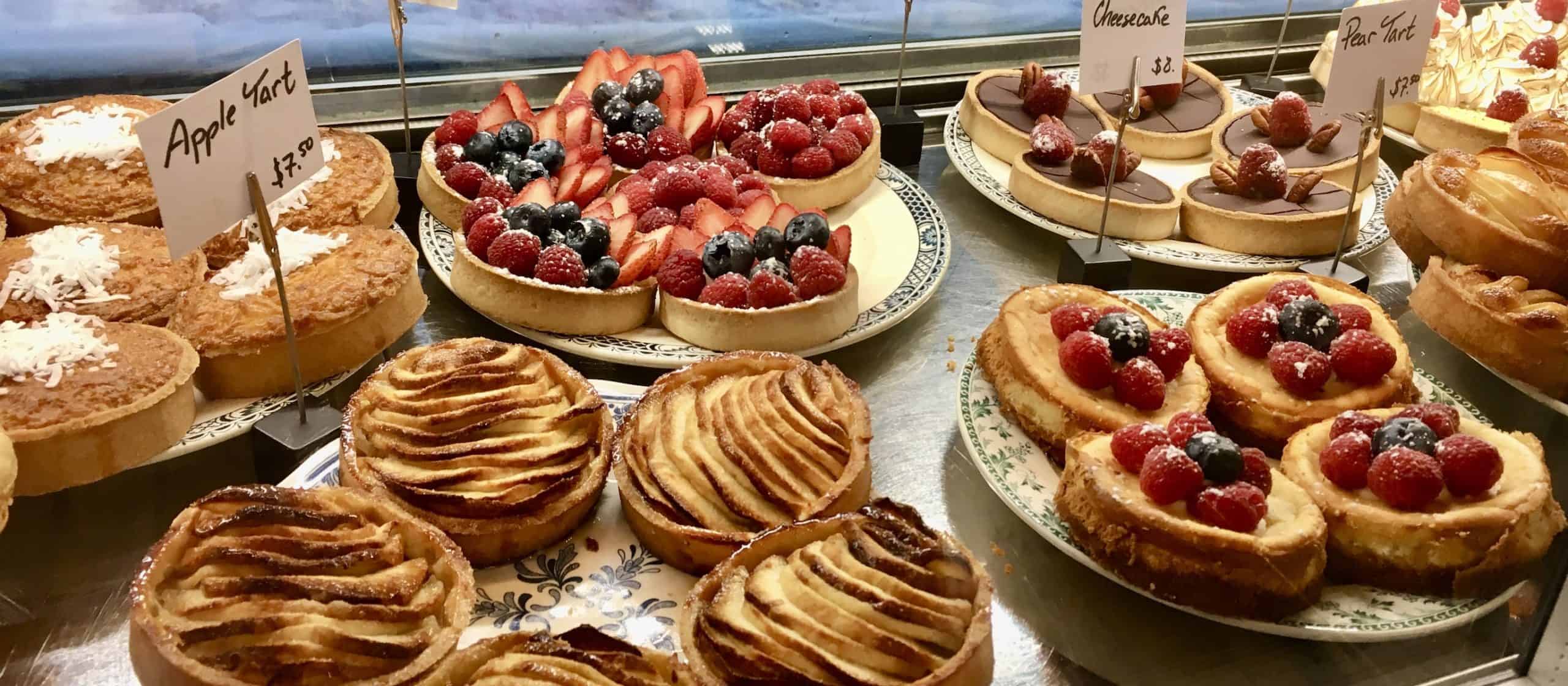 Gluten-free Parisian-style pastries and tarts in a display case, signs say "apple tart $7.50", "cheesecake $8", and "pear tart $7.50"