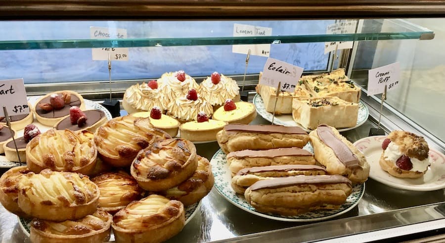 display case with pastries, including tarts, eclairs, mini cheesecakes, chocolate tart, creme puffs - signs say "...tart $7.50", "chocolate tart $7.50", "lemon tart $7.50", "eclair $7.50", "creme puff $7.50", and the sign in the far back right is illegible