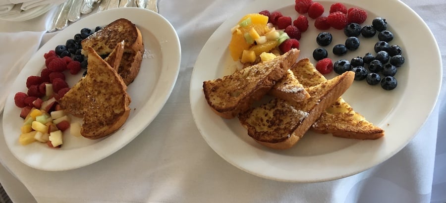 two plates of gluten-free French toast on a white tablecloth, the French taste is cut into triangles and served with a side of raspberries, blueberries, oranges and diced melons