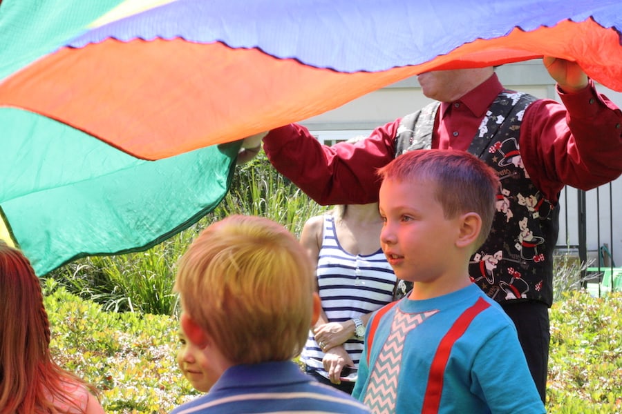 children under a parachute outside, one boy has blue shirt with sewn on tie and suspenders, adult in red shirt and vest standing behind him is holding the parachute