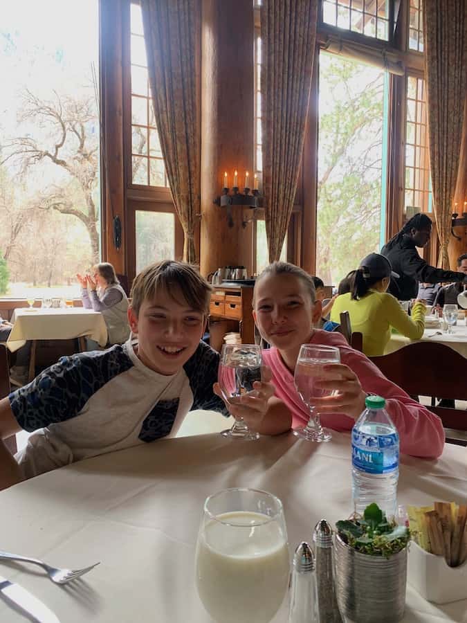 CJ and Miss E holding up water glasses at the Ahwahnee dining room, large windows with Yosemite National Park Views in the background