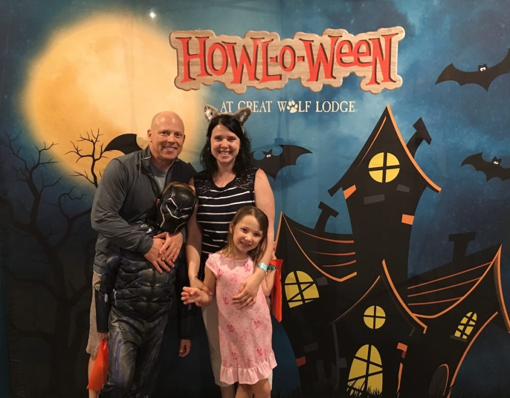 Dave, Heather, CJ in a black panther costume, and Miss E in pink pjs in front of a backdrop of a spooky house with a full moon and the text "Howl-O-Ween, Great Wolf Lodge"