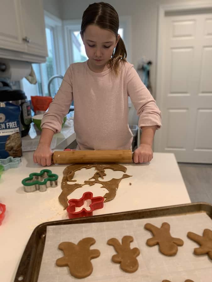 Miss E rolling out part of the gingerbread dough after already cutting out a few shapes in the foreground portion of dough, there are cookie cutters on the counter and a tray of ready to bake gingerbread men on a baking sheet in the foreground