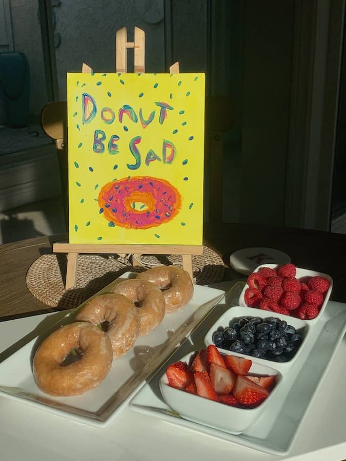 "Donut Be Sad" sign on a yellow background behind a display of Katz glazed donuts and bowls for raspberries, blueberries and strawberries