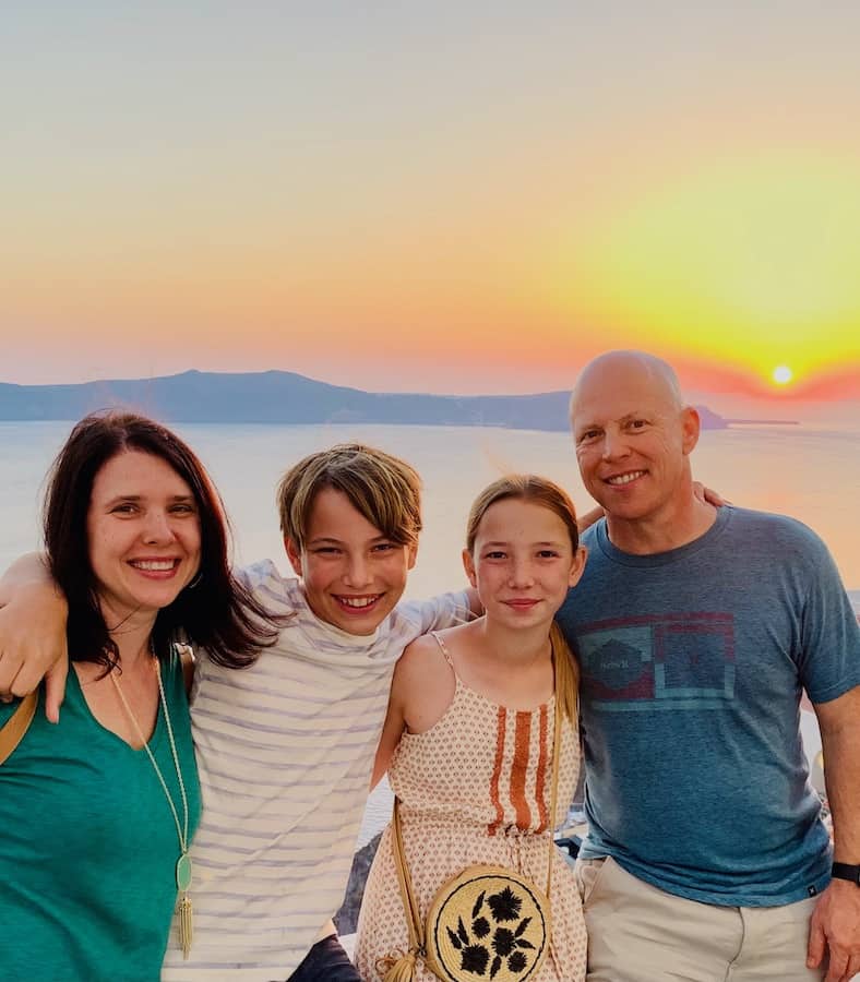 all smiling with arms around each other, Heather in a green t-dress, CJ in a white with blue stripes tee, Miss E in a cream and brown romper, and Dave in a blue t, with the Santorini caldera (volcanic island and water) and sunset in the background