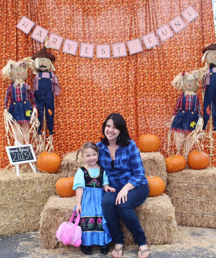 Miss E in a Princess Anna Frozen costume holding a pink pumpkin candy bucket and standing, next to Heather, who is wearing a blue and black flannel shirt and black pants and sitting on a bale of straw. In the background are more bales of straw, orange pumpkins, four scarecrows, and an orange sheet and a banner that reads "harvest fun"