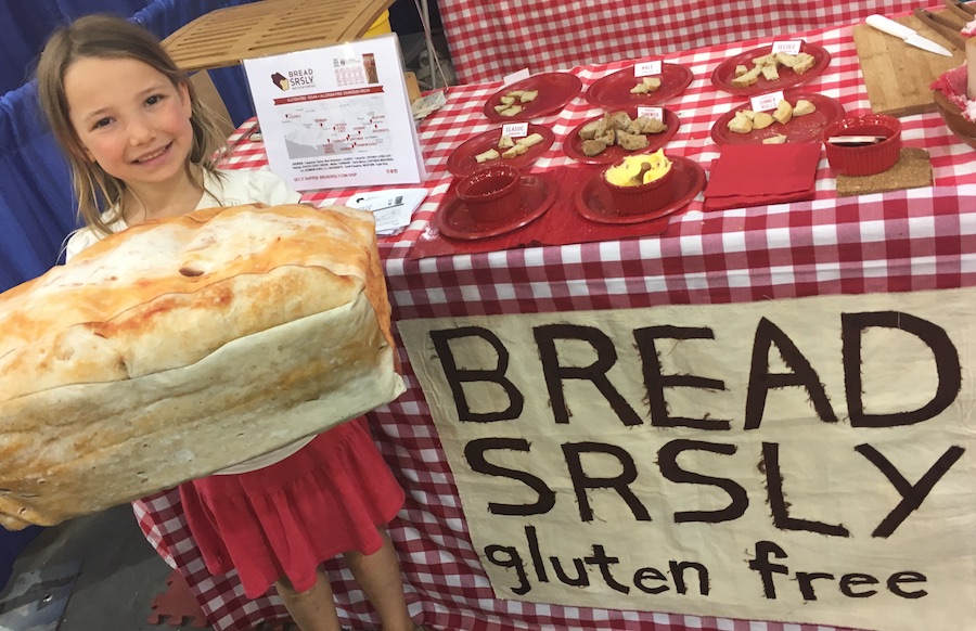a Bread SRSLY booth with the words "BREAD SRSLY gluten free" on the front of the table, a gingham table cloth, with samples on the table, and Miss E in front holding a giant loaf of sourdough bread