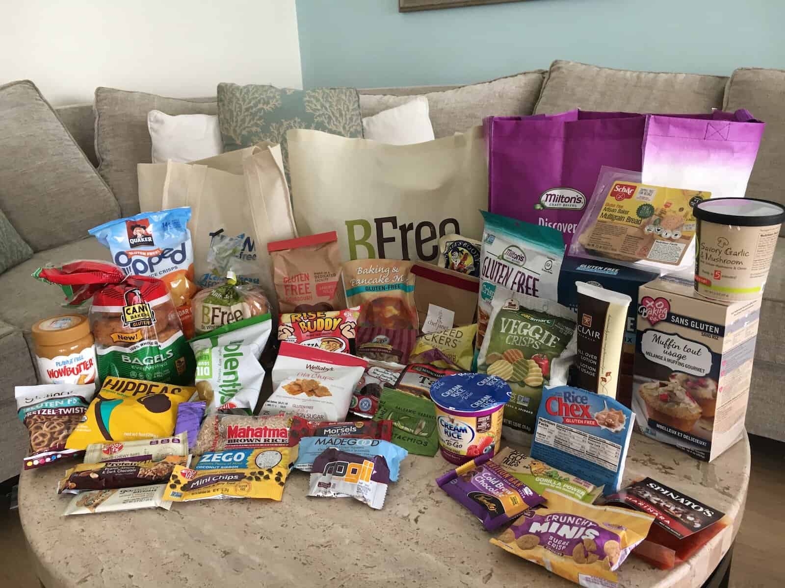 a round travertine coffee table covered with dozens of gluten-free products from bagels to cookies, reusable grocery bags are on the table behind the groceries, a sofa is in the background