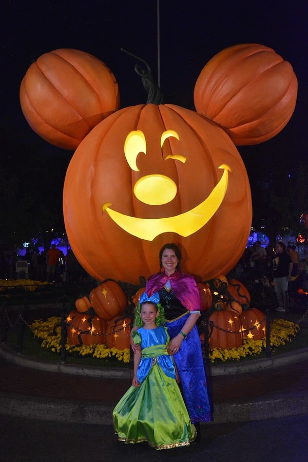 Miss E in a Drizzella costume and Heather in a Princess Anna costume standing in front of a large Mickey Jack-o-lantern