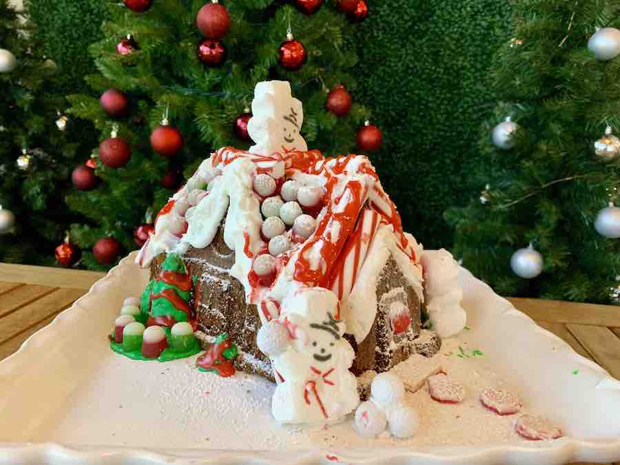 gluten-free gingerbread house covered with frosting and candy, in front of decorated Christmas trees