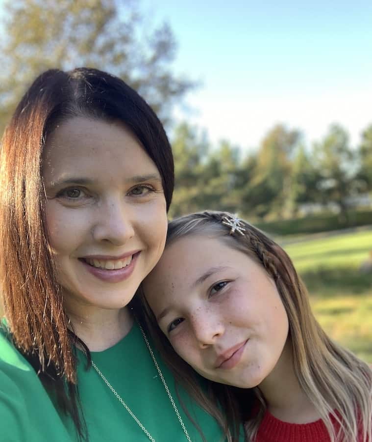 Heather in a green dress with Miss E in a red dress, leaning her head on her mom's shoulder, outside with trees and grass in the background