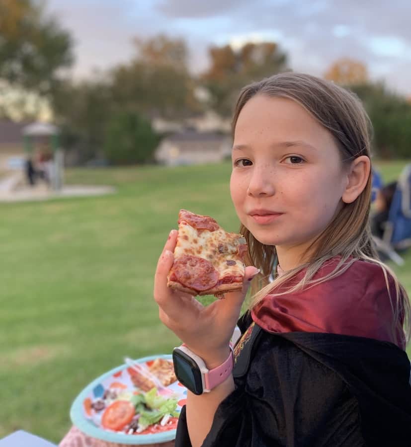 Miss E outside, in a Harry Potter costume, eating a slice of gluten-free pepperoni pizza