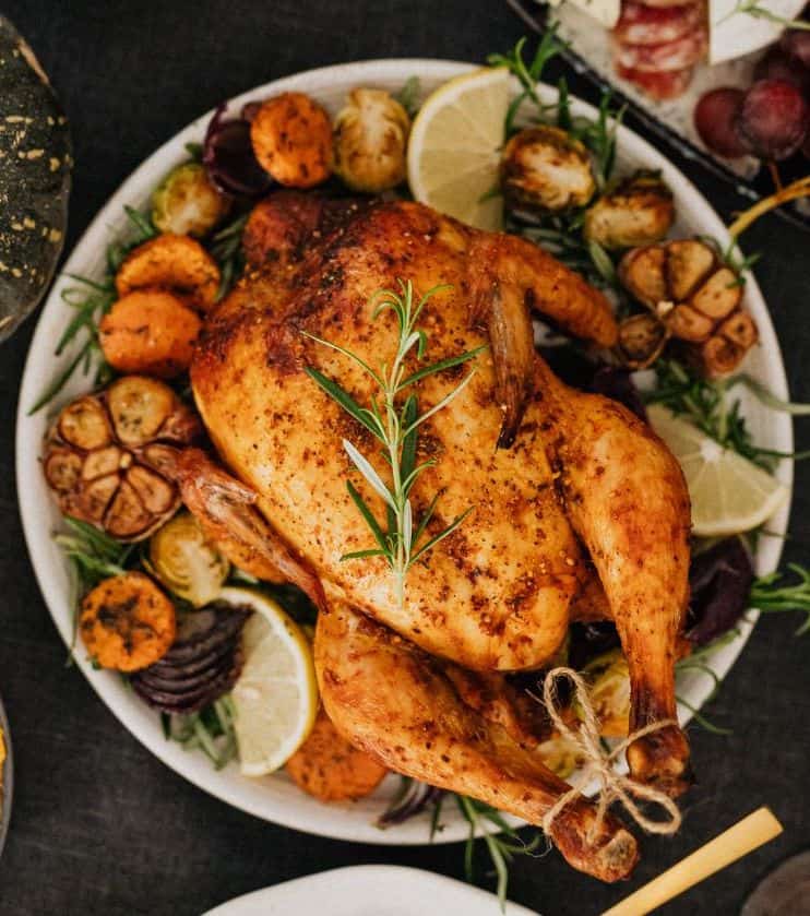 turkey with a sprig of rosemary on top and tied legs, on a bed of vegetables on a round white plate on a table with unidentifiable side dishes