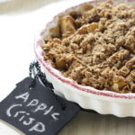 gluten-free apple crisp in a white pie dish with a Burgundy trim, small chalkboard sign on the white, wood table reads: apple crisp
