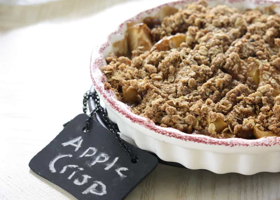 apple crisp in a white pie dish with a Burgundy trim, small chalkboard sign on the white, wood table reads: apple crisp