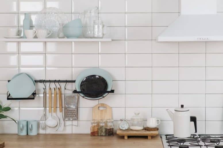 white subway tile, kitchen wall with shelf holding dishes, rack holding dishes and utensils, kitchen counter with jars and mugs, stoves with a white kettle and white, overhead vent