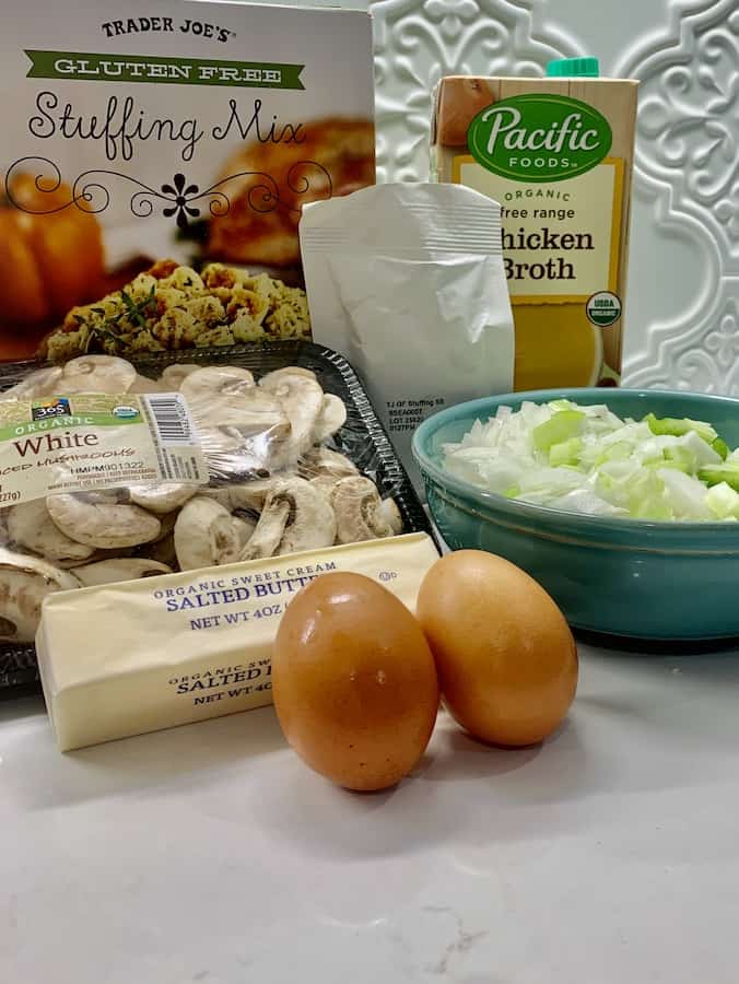 a box of Trader Joe's gluten-free stuffing mix, the white seasoning packet from the stuffing mix, a box of chicken broth, a package of sliced mushrooms, a blue bowl of chopped onions and celery, a wrapped stick of butter and two brown eggs, on a white k