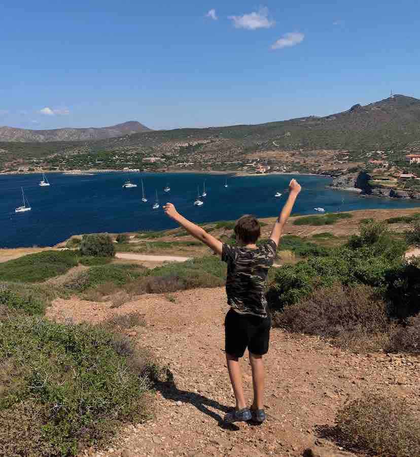 CJ with fists in the air walking on a path above the sea in Cape Sounion