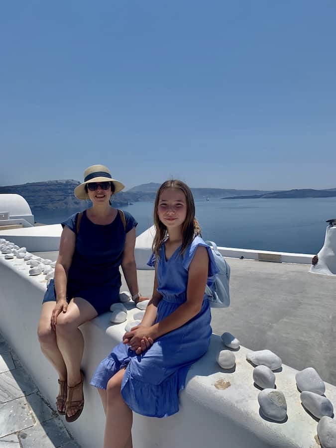 Heather with a hat and Miss E with a backpack, sit-in on a whitewashed ledge in Oia with the blue sea in the background