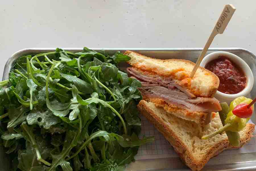 gluten-free mobile order from Pym's test kitchen, a side of arugula, two halves of a sandwich with an "allergy" pick, another pick w/ a pickle and peppers, a cup of marinara sauce for dipping, all on a metal tray on a table
